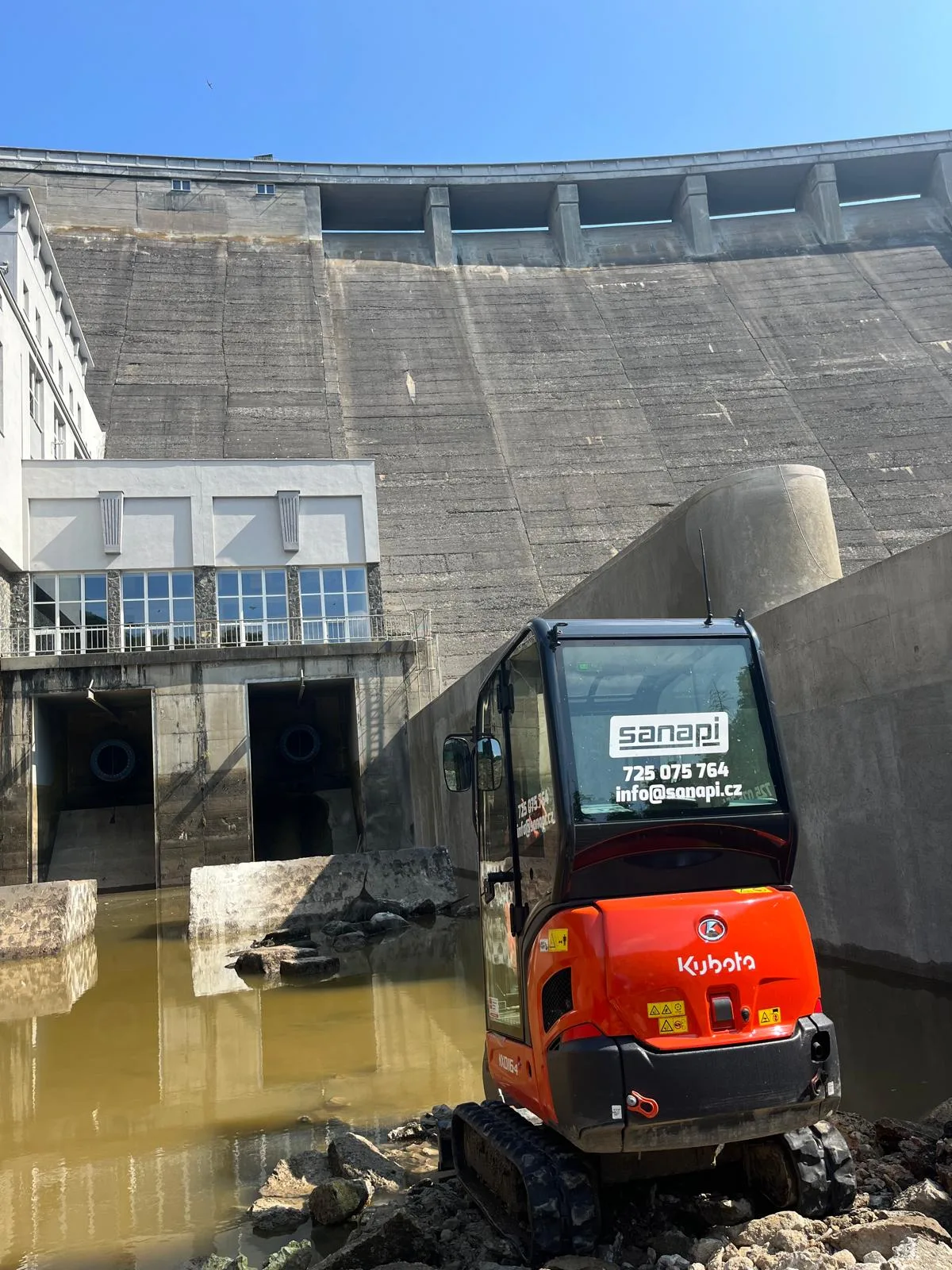 Minirypadlo Kubota s logem SANAPI pod betonovou hrází